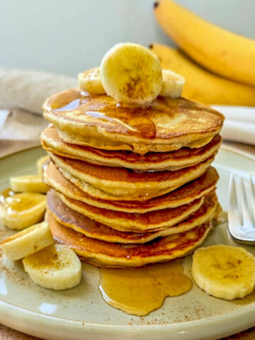 Une pile de pancakes healthy à la banane et sirop d'érable, parsemée de rondelles de banane fraîche, de cannelle servie sur une assiette en grès avec une fourchette.