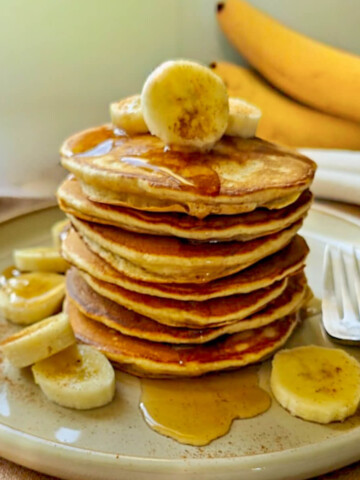 Petit-déjeuner sain : pile de pancakes moelleux à la banane et sirop d'érable, parsemée de rondelles de banane fraîche, de cannelle et de sucre glace, servie sur une assiette en grès avec une fourchette. Recette facile et rapide.