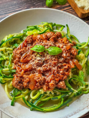 Vue aérienne (flat lay) d'une assiette de zoodles à la bolognaise. Le plat est entouré d'un torchon vichy, de feuilles de basilic frais et d'un petit bol de parmesan râpé.
