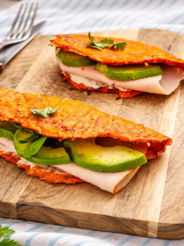 Wraps de carottes faciles et rapides, pliés en deux, garnis de tranches de blanc de dinde, d'avocat frais et d'une base crémeuse, présentés sur une planche à découper avec des couverts en arrière-plan.