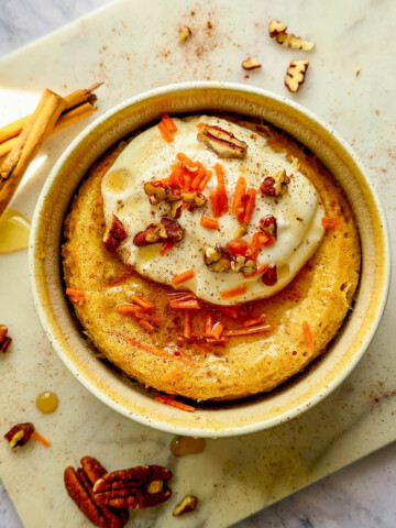 Un bowl cake façon carrot cake dans un bol beige, surmonté de fromage blanc, de carottes râpées et de noix de pécan sur une planche en marbre.