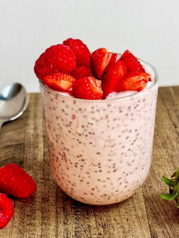 Pudding de chia à la fraise et framboise servi dans un verre transparent sur une planche à découper. Le verre est généreusement garni de fruits rouges frais. Une cuillère et quelques fruits entiers sont disposés en arrière-plan sur le bois rustique.