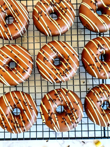 Des donuts au four chocolat cuits au four, disposés sur une grille de refroidissement noire, avec un glaçage brillant au chocolat au lait décoré de zébrures de chocolat blanc et de vermicelles. Un donut sur le côté est entamé pour révéler une texture moelleuse à l'intérieur.