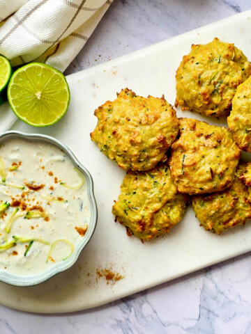 Galettes de courgettes et lentilles corail dorées à l'Airfryer, servies sur une planche blanche avec une sauce au yaourt et des quartiers de citron vert.