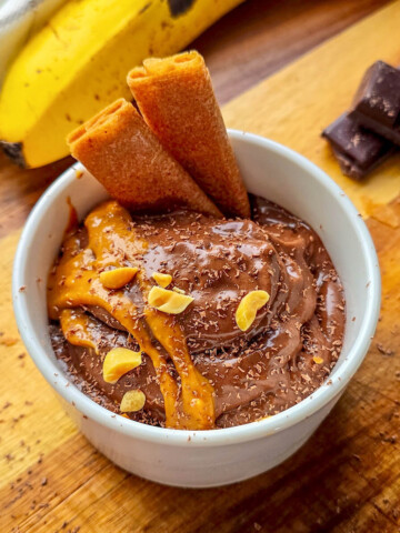 Recette healthy de glace banane chocolat maison sans sorbetière. Bol de nice cream onctueuse nappée de beurre de cacahuètes, parsemée d'éclats de cacahuètes et de copeaux de chocolat, présentée sur une planche en bois.