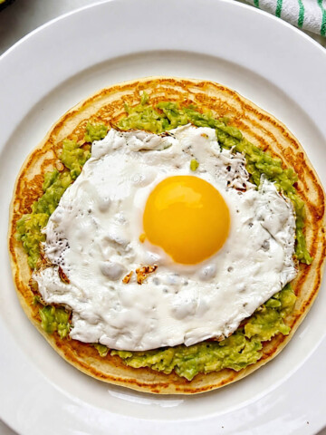 Un pancake salé dans une assiette blanche, garni d'avocat écrasé et d'un oeuf sur le plat, sur un fond blanc marbré, avocat et torchon en arrière plan.