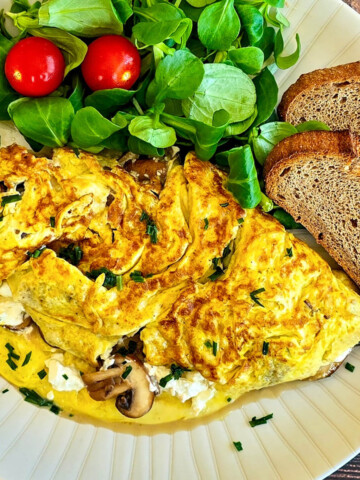 Une omelette aux champignons feta dans une assiette, avec des tranches de pain de campagne et une salade de mâche, sur un fond de bois.