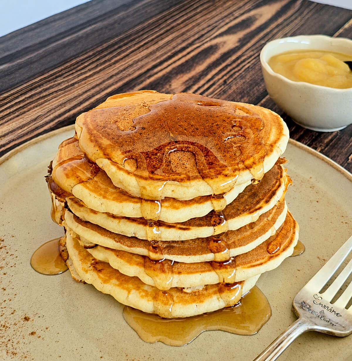 Des pancakes sans oeufs empilés les uns sur les autres, sur une assiette beige, sur un fond de couleur bois, avec du sirop d'érable coulant de chaque côté.