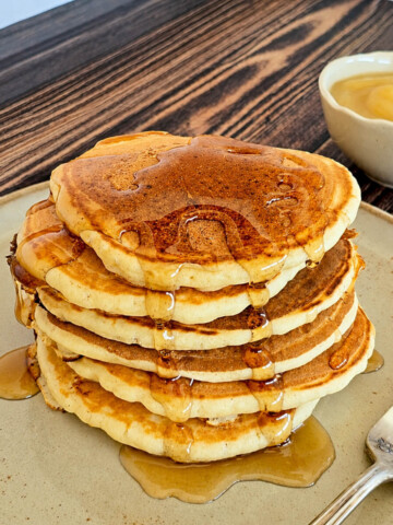 Des pancakes empilés les uns sur les autres, sur une assiette beige, sur un fond de couleur bois, avec du sirop d'érable coulant de chaque côté.
