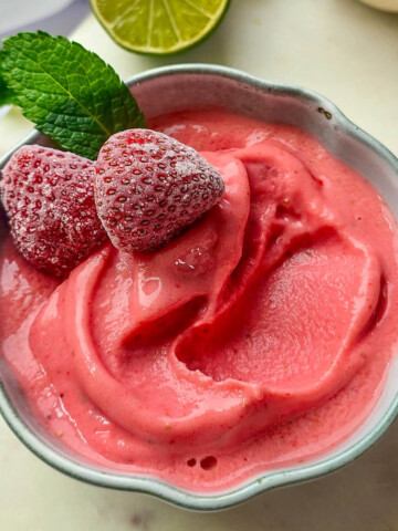 Recette de sorbet fraise maison facile et rapide à faire sans sorbetière. Vue de dessus d'un bol de glace à la fraise onctueuse, décoré de fraises givrées et de menthe fraîche, avec des citrons verts en arrière-plan.