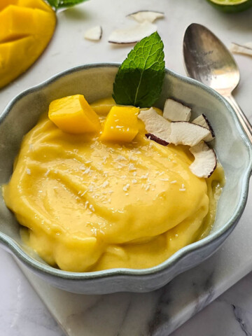 Sorbet mangue maison rapide et rafraîchissant, réalisé sans sorbétière. Présentation du bol sur une planche en marbre avec une cuillère gravée Cuisine & Création et une mangue fraîche coupée à côté.