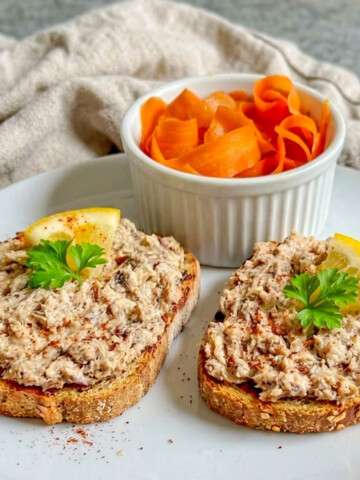 Gros plan de deux tartines croustillantes garnies de rillettes de sardines maison, surmontées d'un quartier de citron frais et de persil, avec un ramequin de tagliatelles de carottes en arrière-plan.