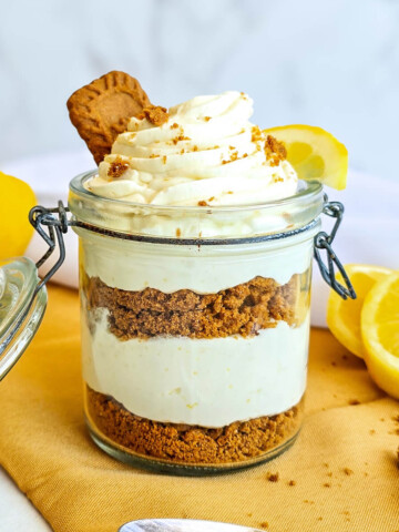 Vue d'ensemble de la verrine citron-speculoos posée sur un tissu jaune. On y voit clairement l'alternance des couches de biscuit brun et de crème blanche. Au premier plan, une cuillère en argent gravée est posée sur la table, et des citrons frais ainsi que des biscuits décorent l'arrière-plan.