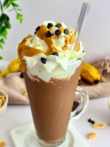 Un milkshake Snickers dans un grand verre avec paille, garni de chantilly, caramel et pépites de chocolat, entouré d'ingrédients.