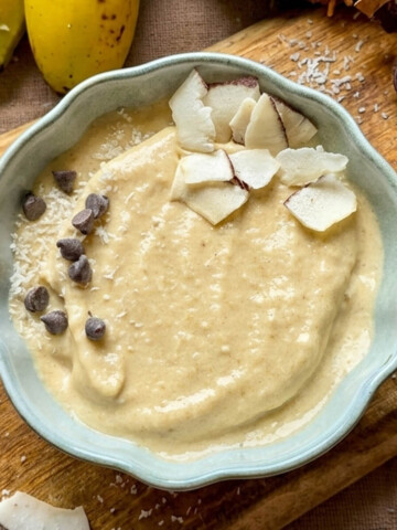 Bol de glace onctueuse banane-coco décoré de copeaux de noix de coco fraîche et de pépites de chocolat noir, posé sur une planche en bois rustique avec deux bananes mûres et une noix de coco entière en arrière-plan.