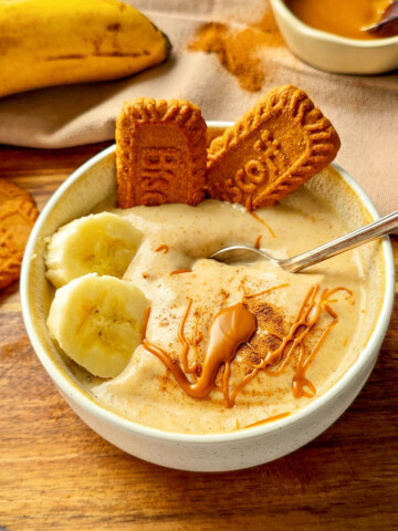 Bol de glace à la banane onctueuse, garni de deux biscuits Biscoff, de rondelles de banane fraîche, d'un filet de beurre de spéculoos et d'une touche de cannelle, présenté sur une planche en bois avec des bananes entières en arrière-plan.
