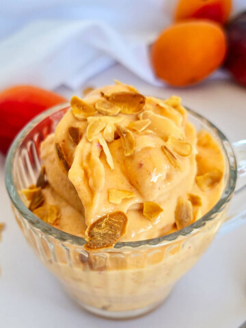 Une coupe en verre givrée remplie d'une glace onctueuse à la pêche et à l'abricot, parsemée d'amandes effilées grillées, avec des fruits frais en arrière-plan.