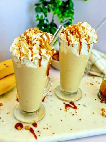 Deux verres de milkshake crémeux au caramel et à la banane sur un plateau en marbre, décorés de crème fouettée, de sauce caramel et de morceaux de banane. À l'arrière-plan, des bananes entières, une petite tasse de sauce caramel et une serviette de table.