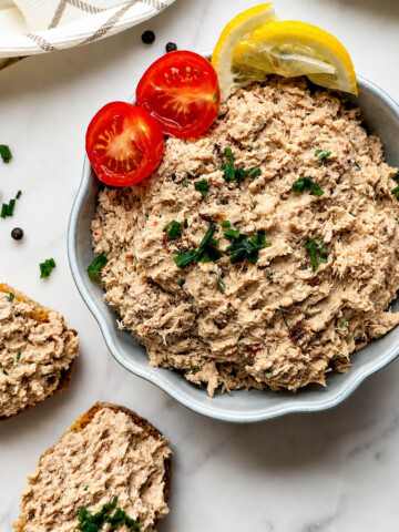 des rillettes de thon faciles et crémeuses dans un bol blanc avec une tomate cerise coupée en deux, et sur des tracnhes de pain, sur un fond blanc marbré,