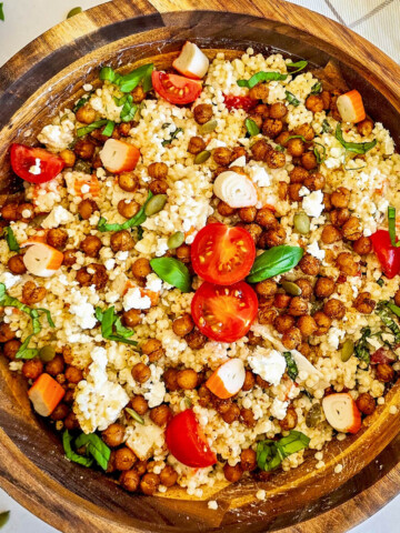 un saladier en bois contenant une salade de perles de blé, pois chiches épicés et rôtis, feta, tomates cerises et herbes fraîches, avec un petit bol de pois chiches rôtis supplémentaires et des graines de courge éparpillées sur une surface claire.