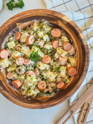 Vue de dessus d'un grand bol en bois rempli d'une salade strasbourgeoise. sur une table blanche, à côté d'un torchon quadrillé gris et blanc et de couverts de service en bois.
