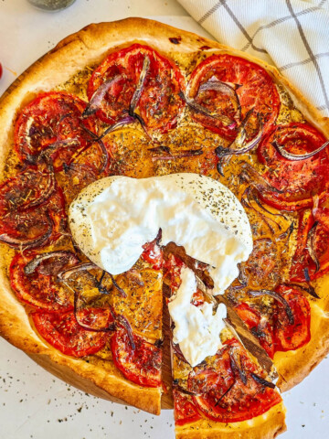 Une part de tarte fine aux tomates et oignons rouges, surmontée de burrata crémeuse, est soulevée au-dessus du plat posé sur une table en marbre blanc.