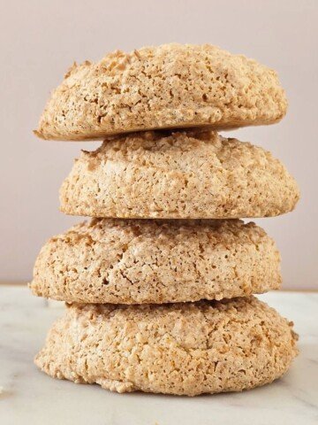 Pile de quatre biscuits à la noix de coco dorés et croustillants, posée sur un plan de travail en marbre blanc devant un fond rose pâle.