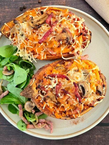 Deux patates douces farcies aux lardons et champignons, gratinées au fromage râpé, servies avec de la mâche dans une assiette beige sur une table en bois foncé.