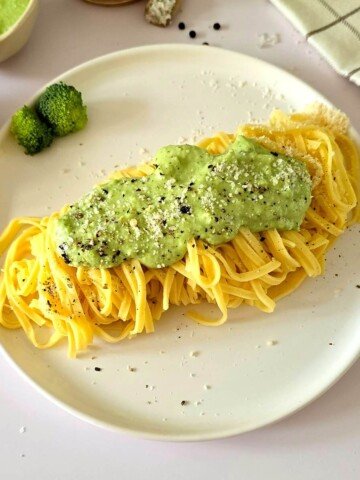 Pâtes au brocoli à la crème sur une assiette blanche, avec du parmesan et de la crème de brocoli sur les bords, le tout sur un fond blanc avec un torchon à carreaux.